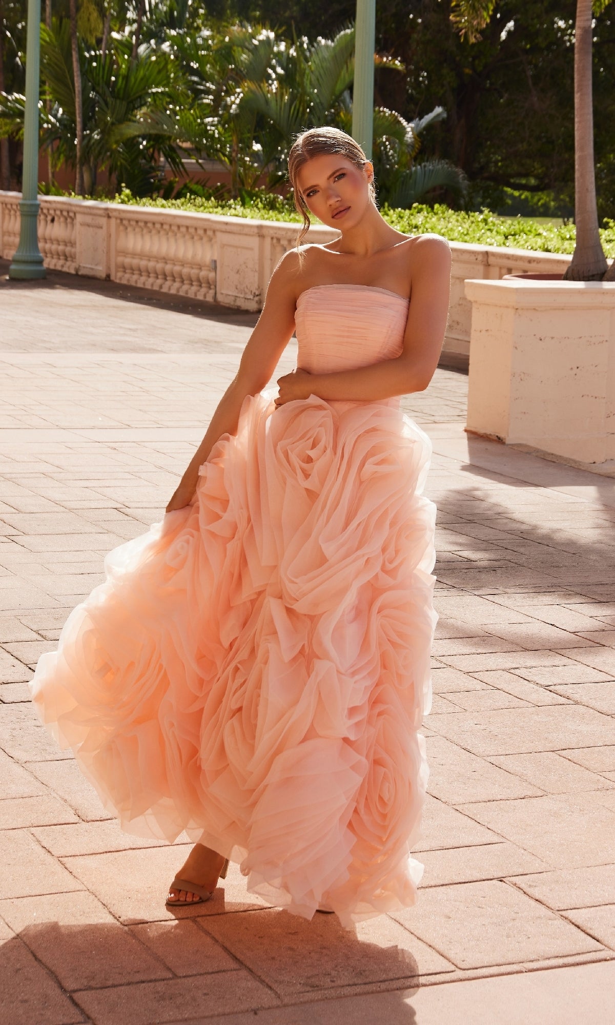 A woman stands outdoors on a sunlit walkway, greenery behind her, wearing the Nox Anabel JQ1037 Strapless Peach Prom Ball Gown with a voluminous ruffled skirt.